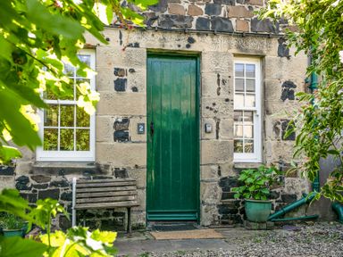 A stone house facade with a green door two white windows a wooden bench and plants at Elliot Cottage in Bamburgh