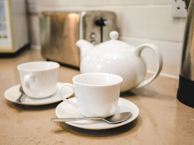 A kitchen countertop with two white teacups on saucers with spoons and a white teapot with a stainless steel toaster in the background at Elliot Cottage in Bamburgh