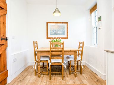 A dining area with a wooden table and four chairs with white cushions a hanging light a window and a framed picture on the wall at Elliot Cottage in Bamburgh