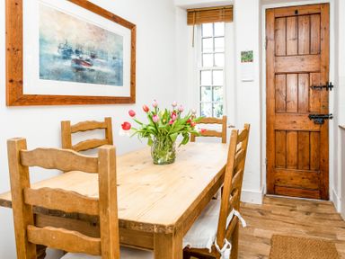 A dining area with a wooden table and four chairs a vase with flowers on the table a wooden door and a window with blinds at Elliot Cottage in Bamburgh