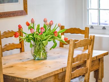 A wooden dining table with a glass vase of tulips and wooden chairs near a window at Elliot Cottage in Bamburgh