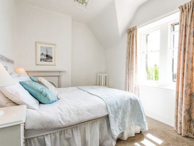 A bedroom with a bed with blue pillows and a blanket near a window with patterned curtains at Elliot Cottage in Bamburgh