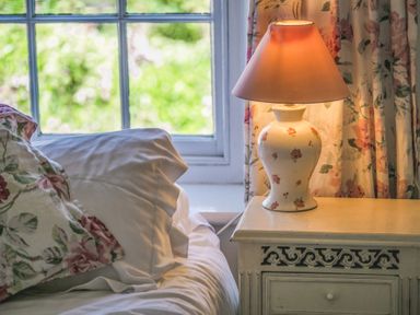 A bedside table with a floral lamp next to a bed with floral pillows and a window in the background at Elliot Cottage in Bamburgh
