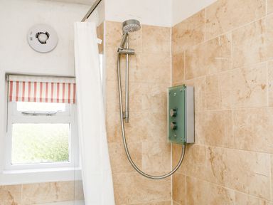 A shower area with a showerhead and control panel next to a window with a striped blind and a white shower curtain at Elliot Cottage in Bamburgh