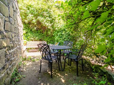 An outdoor seating area with a metal table and four chairs next to a stone wall and surrounded by greenery at Elliot Cottage in Bamburgh