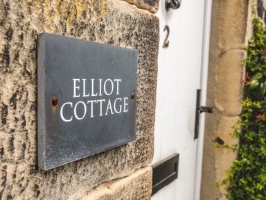 A stone wall with a sign reading Elliot Cottage next to a white door with the number 2 and a black handle at Elliot Cottage in Bamburgh