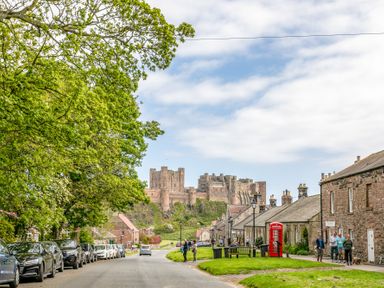 A street with cars parked along the left and people walking near stone buildings with a red telephone box and a large castle on a hill in the background at Elliot Cottage in Bamburgh