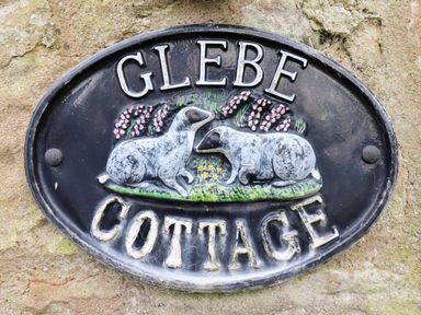 A black oval sign with two white sheep and flowers reading Glebe Cottage attached to a stone wall