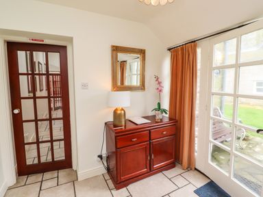A hallway with a wooden cabinet holding a lamp and flower next to glass doors with orange curtains at Glebe Cottage in Bamburgh