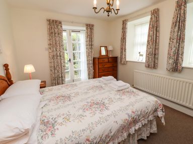 A bedroom with a bed with floral bedding a wooden chest of drawers bedside table lamps and floral curtains at Glebe Cottage in Bamburgh