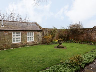 A stone wall garden with grass and flowers and a small stone building with windows at Glebe Cottage in Bamburgh