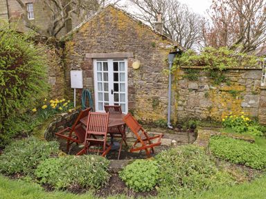 An outdoor patio area with wooden table and chairs against stone walls and plants at Glebe Cottage in Bamburgh