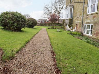 A gravel pathway with bushes and trees beside a stone building with white metal chairs and tables on grass at Glebe Cottage in Bamburgh