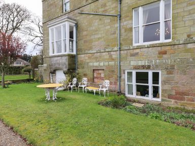 An outdoor area with a stone building wall three white metal chairs one white metal table and a green lawn at Glebe House in Bamburgh