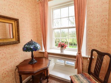 A corner of a room with a wooden table, stained glass lamp, vase with flowers on windowsill, framed mirror, and wooden chair at Glebe House in Bamburgh
