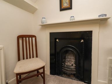 A corner of a room with a wooden chair and a fireplace with a white mantel and decorative items on top at Glebe House Bamburgh