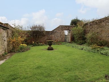 A garden with green grass flower beds and a stone wall with a metal gate at Glebe House in Bamburgh