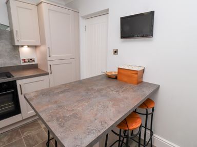 A kitchen with a countertop island and two stools with wooden seats a small wall-mounted television and kitchen cabinets at Bay View in Amble