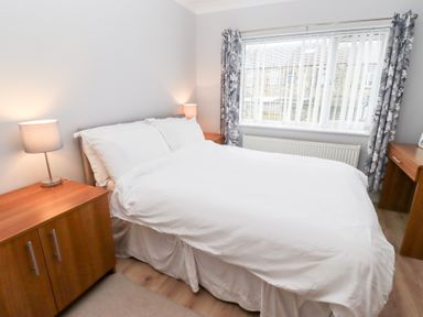 A bedroom with a double bed white bedding wooden bedside tables table lamps window with vertical blinds and floral curtains at Bay View in Amble