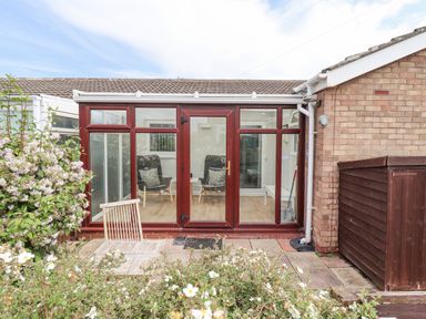 A glass conservatory with chairs inside and a small wooden table chair outside with flowers in the garden at Bedwine Cottage in Beadnell