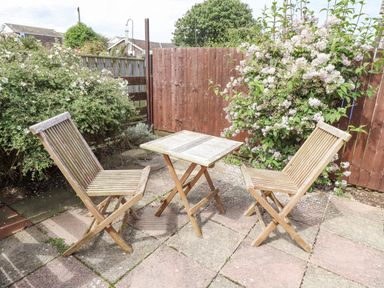 An outdoor patio with two wooden chairs and a wooden table surrounded by flowering bushes and a wooden fence at Bedwine Cottage in Beadnell
