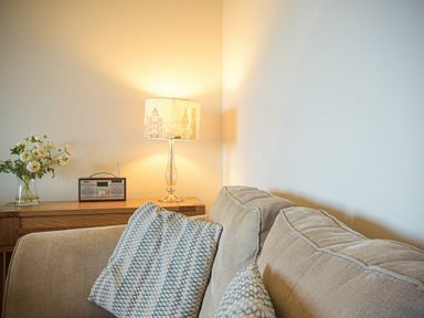 A beige sofa with patterned cushions next to a wooden side table with a lamp radio and flowers at Bellshill Cottage in Belford