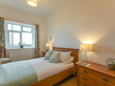 A bedroom with a wooden bed, bedside tables with lamps, a window with floral curtains, and a wooden chest of drawers at Bellshill Cottage in Belford