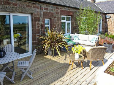 An outdoor deck with a table and chairs a sofa with cushions a small table with flowers and several potted plants at Bellshill Cottage in Belford