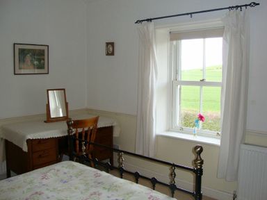 A bedroom with a bed frame, a wooden desk and chair, a mirror, a window with curtains and a flower on the windowsill at Briar Cottage in Town Yetholm and Kirk Yetholm