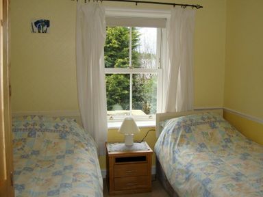 A bedroom with two single beds a wooden nightstand with a lamp and a window with white curtains at Briar Cottage Town Yetholm and Kirk Yetholm
