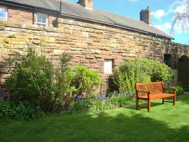 A garden with bushes flowers and a wooden bench next to a stone wall at Briar Cottage in Town Yetholm and Kirk Yetholm