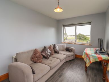 A room with a beige sofa with pillows a table with a checkered cloth and a window showing outdoor greenery at Budle Bay Cottage Bamburgh