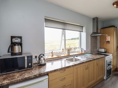 A kitchen with a microwave coffee maker kettle sink stove and wooden cabinets below a window at Budle Bay Cottage in Bamburgh