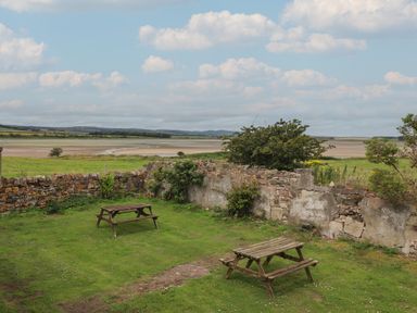 A garden with two wooden picnic tables and a stone wall overlooking fields and water at Budle Bay Cottage in Bamburgh
