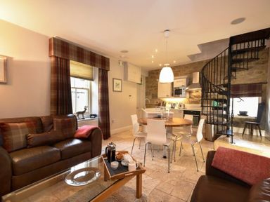 An open living area with leather sofas a glass coffee table a round dining table with chairs a kitchen and a spiral staircase at Coquet Cottage Warkworth