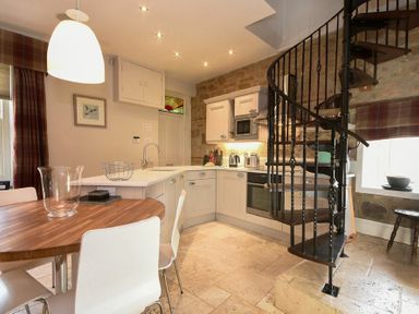 A kitchen with white cabinets a wooden dining table and a black spiral staircase at Coquet Cottage Warkworth