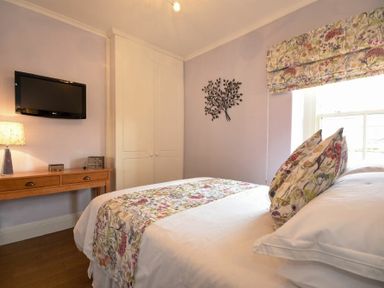 A bedroom with a bed floral pillows and runner a wooden desk a wall mounted television and a wardrobe at Coquet Cottage Warkworth