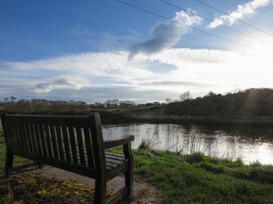 A wooden bench facing a river with grassy banks and trees under a partly cloudy sky at Coquet Cottage Warkworth