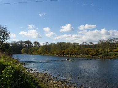 A river with a rocky shore and trees along the banks under a blue sky with clouds at Coquet Cottage Warkworth