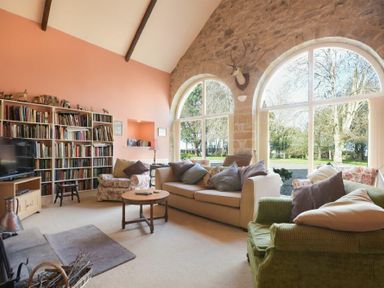 A living room with a bookcase television sofas armchairs wooden coffee table and large arched windows at Craster Tower Coach House Craster