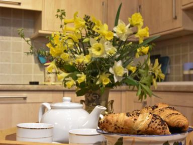 A kitchen counter with a bouquet of yellow and white flowers a white teapot two teacups and a plate of croissants at Craster Tower Coach House in Craster