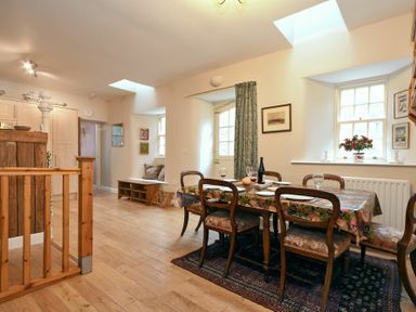 A dining area with a wooden table and six chairs next to windows and a wooden stair railing at Craster Tower Coach House in Craster