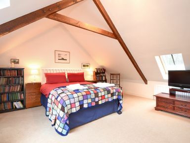 A bedroom with a double bed covered with a patchwork quilt two red pillows bookshelves a wooden chest of drawers a wooden chair a television and a skylight window at Craster Tower Coach House in Craster
