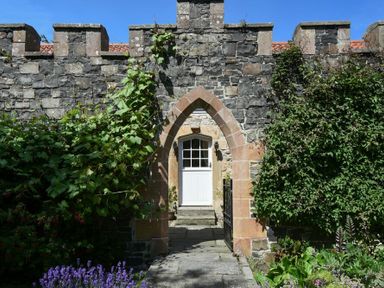 A stone arched doorway in a medieval stone wall with a white door behind it and plants growing on both sides at Craster Tower Coach House in Craster