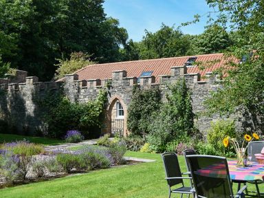 A garden with outdoor chairs and table with flowers beside a stone wall and building with tiled roof at Craster Tower Coach House in Craster