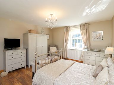 A bedroom with a metal bed frame a dresser with a television a wardrobe an armchair and a window with curtains at Dray Cottage in Warkworth