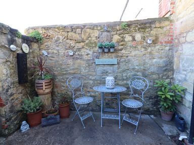 A small outdoor patio with a stone wall two metal chairs a metal table with a lantern and potted plants at Dray Cottage in Warkworth