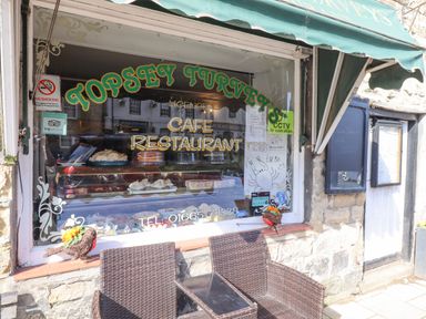 The exterior of Topsey Turvey cafe restaurant with a window display of cakes and a seating area with two brown wicker chairs and a small table at Dray Cottage in Warkworth