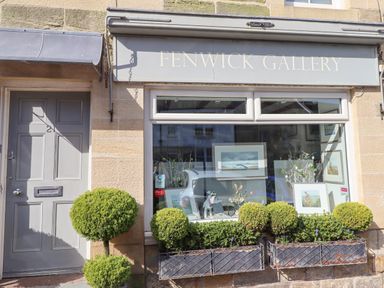 A storefront with framed art in the window and small spherical bushes in planters below at Fenwick Gallery in Warkworth