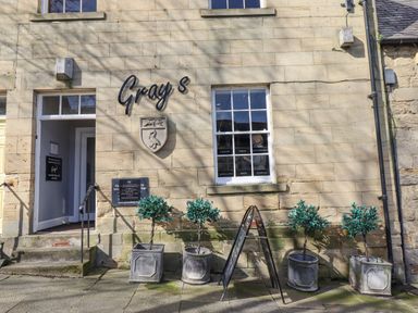 The exterior of a stone building with a door open to a shop named Grays with four potted plants and a sidewalk sign in front
